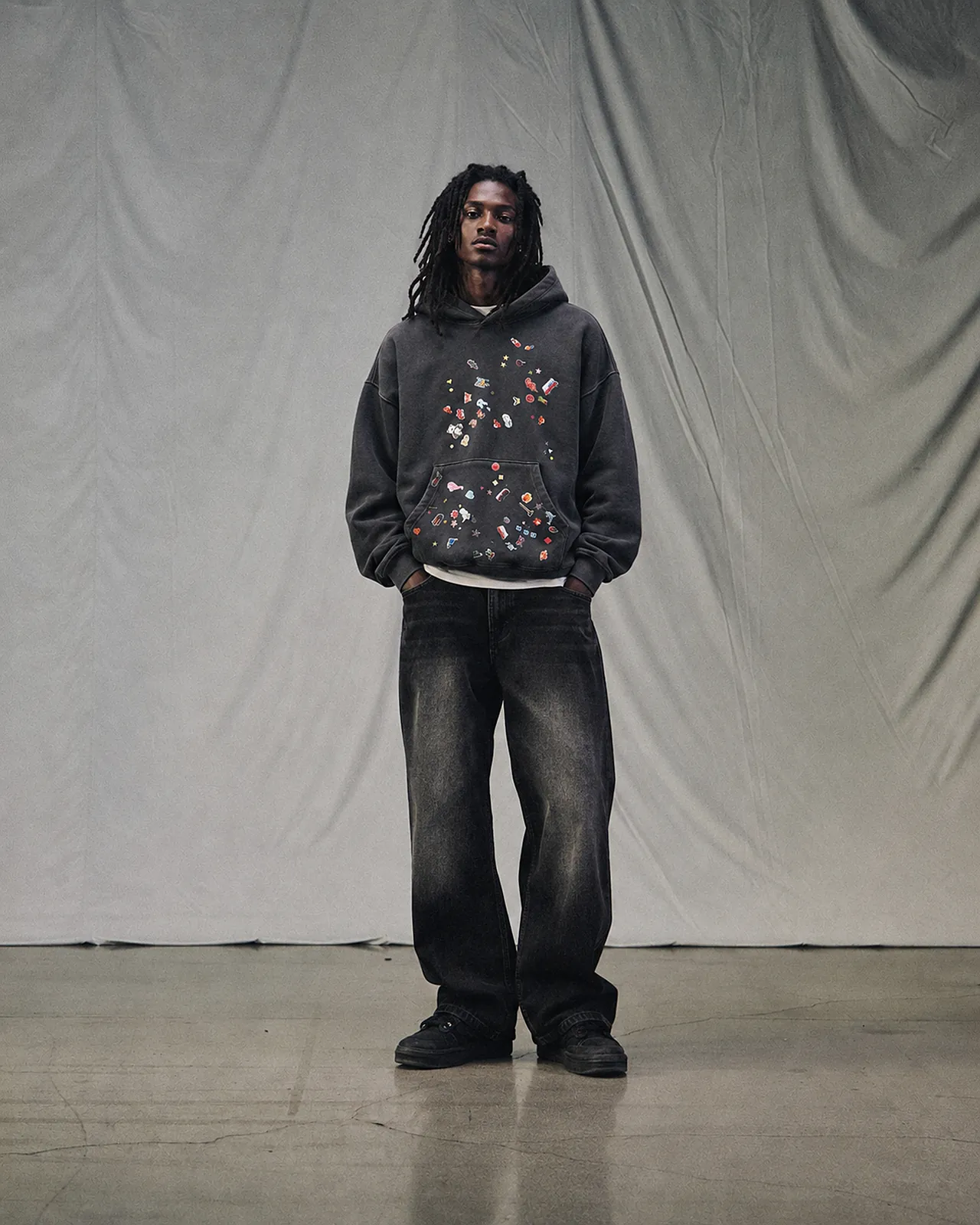 Model with long locs in The Hoodie II and The Americana Washed Jeans VI, standing in front of a white draped cloth
