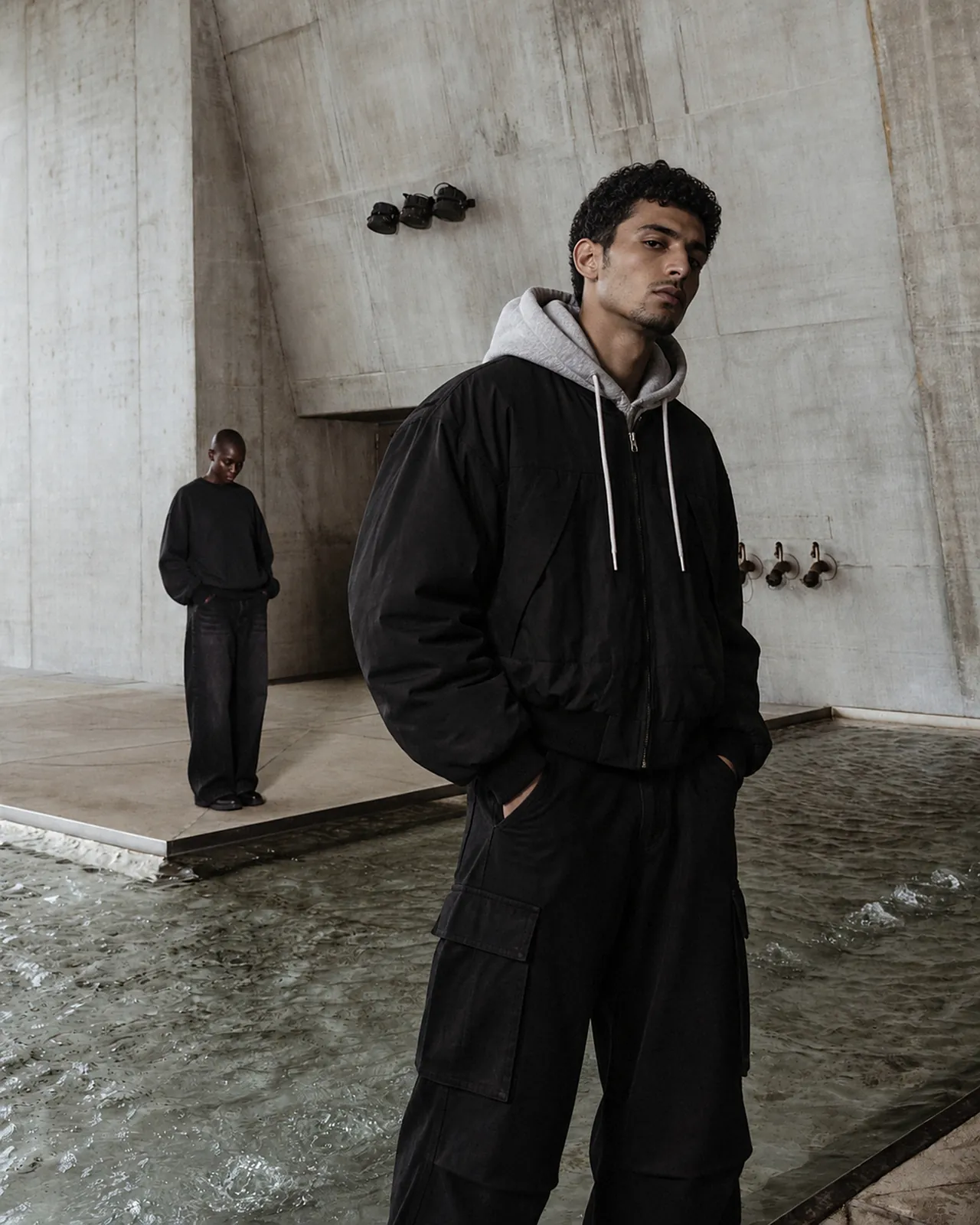 Model in The Layered Jacket and The Street Washed Cargo Trousers in a concrete hall, a second model behind in The Washed Hoodie II and The Street Jeans